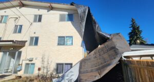 Stettler Wind Storm Tears Roof Off Apartment Building Two-story beige apartment building with a dark tarp-covered collapse in the yard against a clear blue sky.