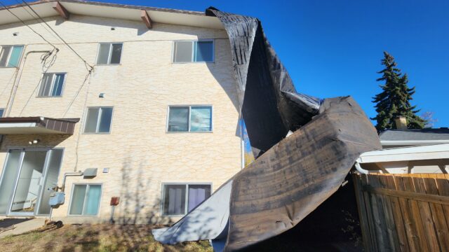 Two-story beige apartment building with a dark tarp-covered collapse in the yard against a clear blue sky.