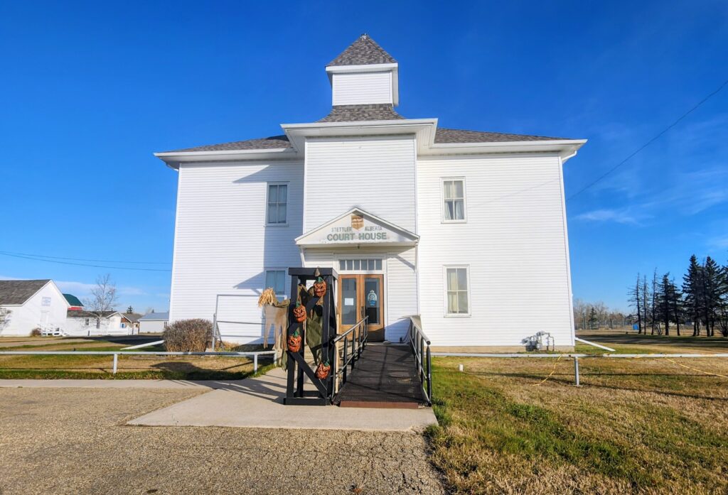 Front view of a white courthouse with Halloween decorations—pumpkins and straw figures—along the railing at the entrance under a blue sky.