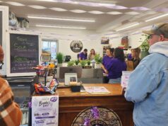 Jacked-Up Coffee Hosts Fundraiser For Stettler Resident’s Medical Expenses People line up at a cafe counter with a chalkboard menu on the left and purple streamers overhead inside a cozy shop. Behind the counter, staff prepare orders while customers wait.