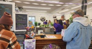 Jacked-Up Coffee Hosts Fundraiser For Stettler Resident’s Medical Expenses People line up at a cafe counter with a chalkboard menu on the left and purple streamers overhead inside a cozy shop. Behind the counter, staff prepare orders while customers wait.