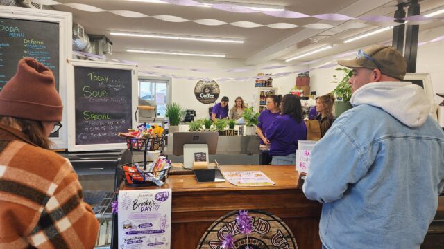 People line up at a cafe counter with a chalkboard menu on the left and purple streamers overhead inside a cozy shop. Behind the counter, staff prepare orders while customers wait.