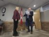 James Nibourg Elected Reeve Of County Of Stettler Two people in a ceremony: a man hands a plaque to a woman in a burgundy blazer in front of flags inside a meeting room.