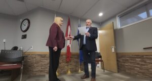 James Nibourg Elected Reeve Of County Of Stettler Two people in a ceremony: a man hands a plaque to a woman in a burgundy blazer in front of flags inside a meeting room.