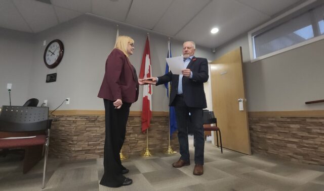 20261027_101011 Two people in a ceremony: a man hands a plaque to a woman in a burgundy blazer in front of flags inside a meeting room.