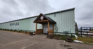 2026 Remembrance Day Service Moves To Stettler Agri-Plex Green corrugated metal warehouse with a wooden entry canopy and an 'AGRIPLEX' sign on the side under a cloudy sky.