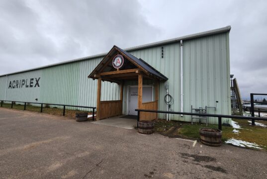 2026 Remembrance Day Service Moves To Stettler Agri-Plex Green corrugated metal warehouse with a wooden entry canopy and an 'AGRIPLEX' sign on the side under a cloudy sky.