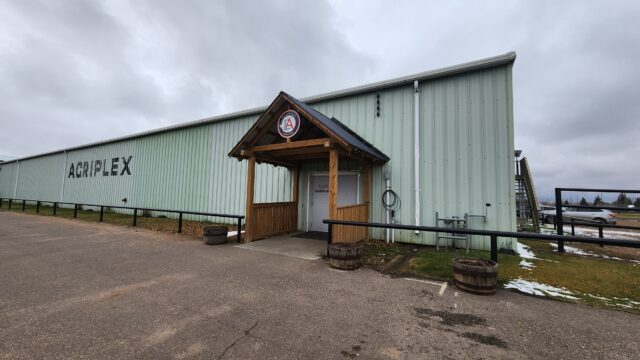 Green corrugated metal warehouse with a wooden entry canopy and an 'AGRIPLEX' sign on the side under a cloudy sky.