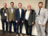 Town of Stettler Presses Province On Policing Cost Disparities At Municipal Convention Five men in business attire wearing conference badges pose together against a light wall, from left to right: casual jacket, brown blazer, navy suit, gray blazer, light gray suit.