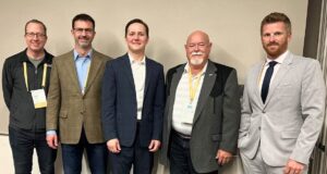 Town of Stettler Presses Province On Policing Cost Disparities At Municipal Convention Five men in business attire wearing conference badges pose together against a light wall, from left to right: casual jacket, brown blazer, navy suit, gray blazer, light gray suit.