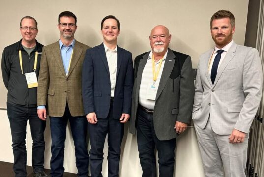 Town of Stettler Presses Province On Policing Cost Disparities At Municipal Convention Five men in business attire wearing conference badges pose together against a light wall, from left to right: casual jacket, brown blazer, navy suit, gray blazer, light gray suit.