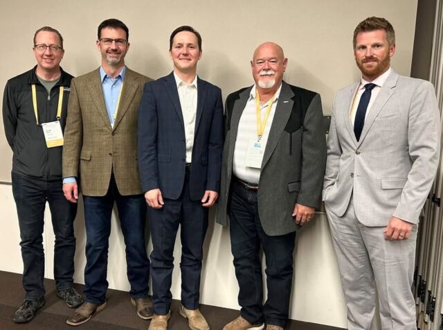Five men in business attire wearing conference badges pose together against a light wall, from left to right: casual jacket, brown blazer, navy suit, gray blazer, light gray suit.