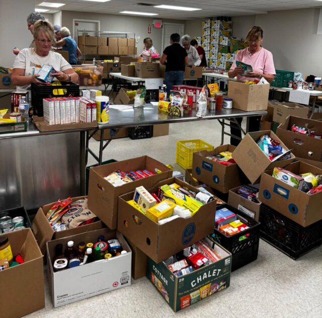 843117033_1414642000790088_3759156947894137670_n Volunteers sorting and packing canned goods and boxed foods at a busy food pantry workstation.