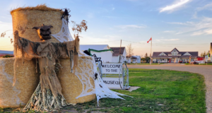 Stettler Booseum 2026: Haunted House Opens At The Museum Courthouse A straw scarecrow figure on a large hay bale with tattered cloth, near a 'Welcome to the Museum' sign and decorative white web, in a rural town during sunset.