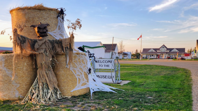 A straw scarecrow figure on a large hay bale with tattered cloth, near a 'Welcome to the Museum' sign and decorative white web, in a rural town during sunset.