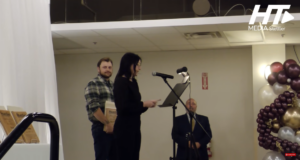 Stettler Awards Gala Celebrates Local Citizens & Businesses Woman speaks at a microphone at a podium on a stage; a man in plaid stands with award plaques behind her, festive balloons to the right.