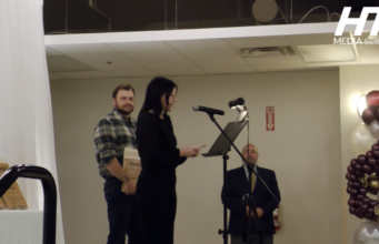 Stettler Awards Gala Celebrates Local Citizens & Businesses Woman speaks at a microphone at a podium on a stage; a man in plaid stands with award plaques behind her, festive balloons to the right.