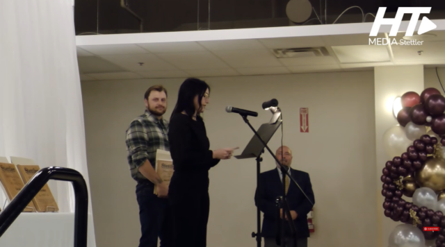 Screenshot 2026-10-27 150541 Woman speaks at a microphone at a podium on a stage; a man in plaid stands with award plaques behind her, festive balloons to the right.