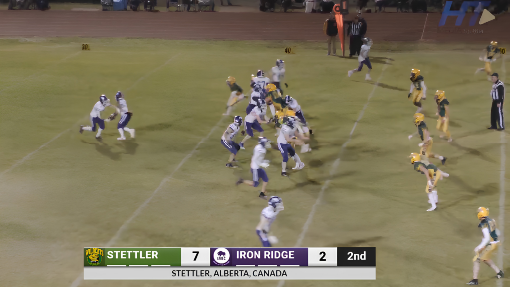 Action shot from a high school football game: Stettler leads Iron Ridge 7-2 in the 2nd quarter as players clash near the line of scrimmage on a green field under stadium lights.