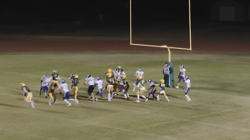 Nighttime football game with players sprinting toward the end zone near a goalpost on a lit field.