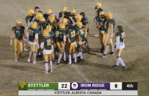 Stettler Wildcats Win First Home Game Since 2022 Football players in green and gold huddle on a dusty field as a scoreboard shows Stettler 22, Iron Ridge 8 in the 4th quarter (Stettler, Alberta, Canada). -Informative image conveying team celebration and score.