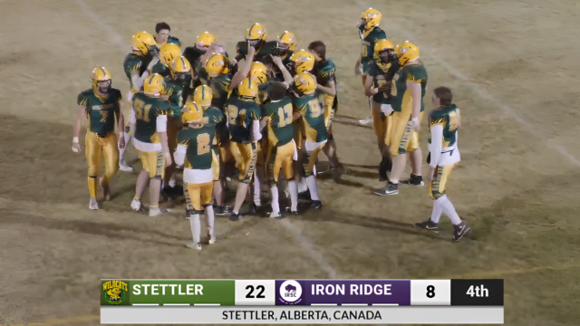 Wildcats 3 Football players in green and gold huddle on a dusty field as a scoreboard shows Stettler 22, Iron Ridge 8 in the 4th quarter (Stettler, Alberta, Canada). -Informative image conveying team celebration and score.
