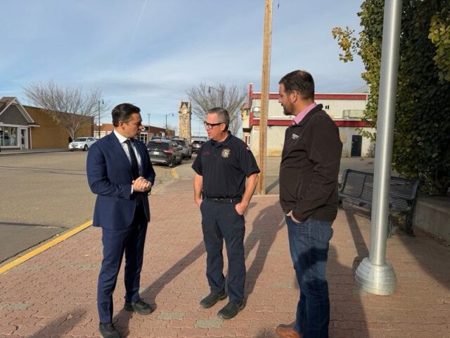 Three men stand on a brick sidewalk in a small‑town street, conversing: a man in a navy suit, a police officer, and a casually dressed man.