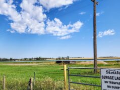 Town of Stettler Awards Lagoon Desludging Project Rural scene with a tall wooden utility pole beside a gate and a sign reading 'Town of Stettler Sewage Lagoons No Trespassing No Dumping' against a blue sky and grassy field.