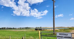Town of Stettler Awards Lagoon Desludging Project Rural scene with a tall wooden utility pole beside a gate and a sign reading 'Town of Stettler Sewage Lagoons No Trespassing No Dumping' against a blue sky and grassy field.
