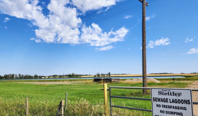 original_12f106c3-4462-44b3-a9b1-c97deda42f53_20250826_132558 Rural scene with a tall wooden utility pole beside a gate and a sign reading 'Town of Stettler Sewage Lagoons No Trespassing No Dumping' against a blue sky and grassy field.